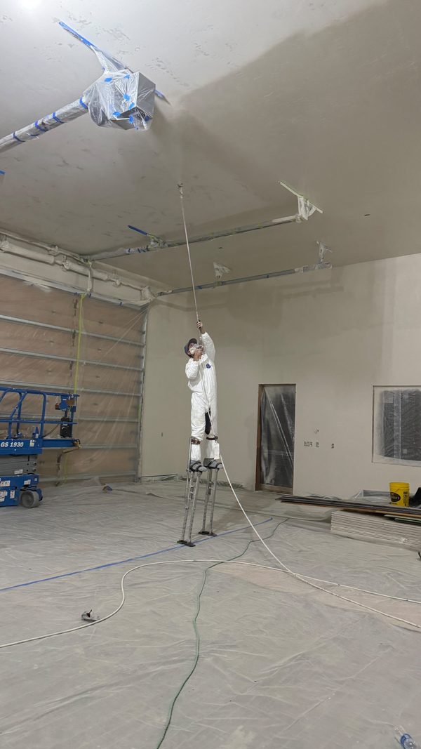 A worker in coveralls spraying the walls of an empty commercial room
